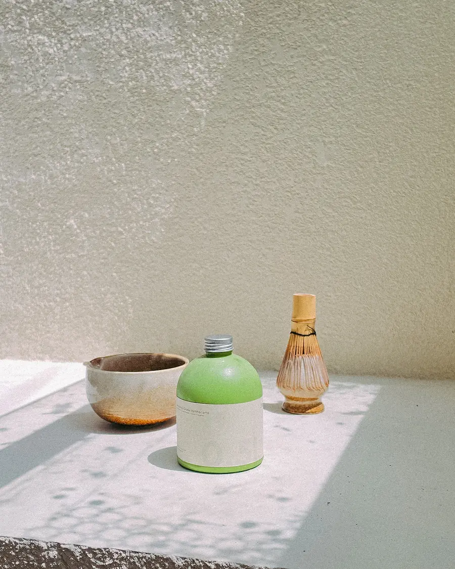 A bottle of Joycha matcha beside a matcha bowl and bamboo whisk in the sun